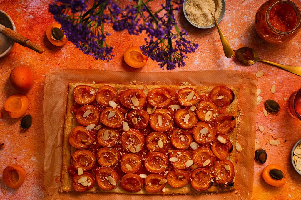Apricot puff pastry tart with apricot halves and flaked almonds on orange background.