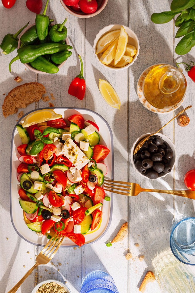 Greek salad in the sunlight on the wooded table with drinks, olives and peppers.