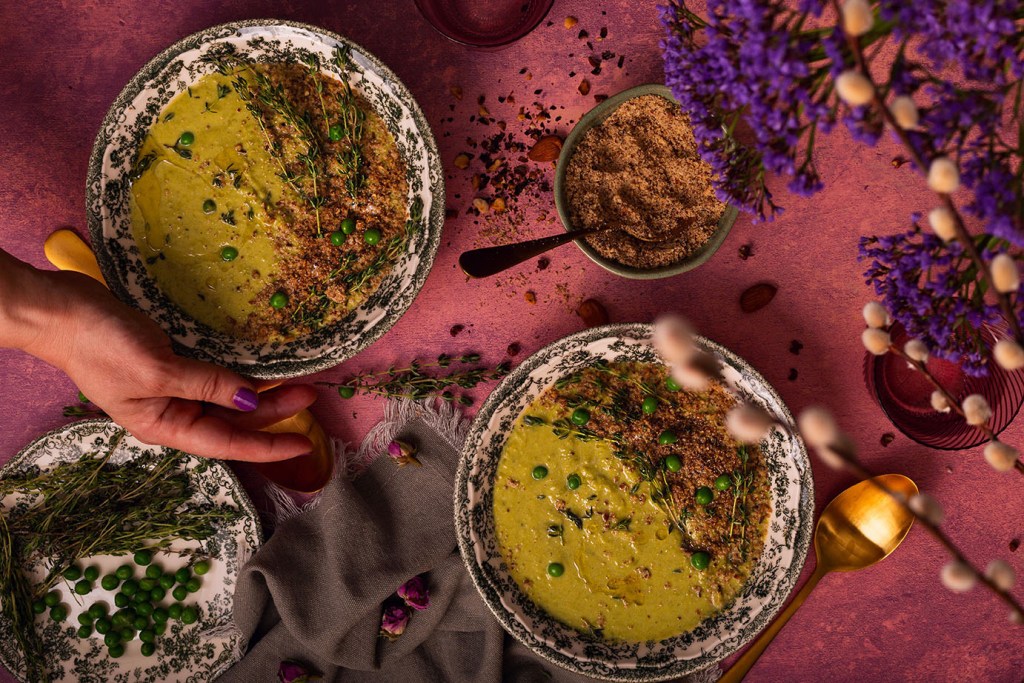 Creamy vegan Pea and Leek Soup with coconut milk, fresh thyme, and dukkah sprinkle in the bowls on the table with hand in frame.